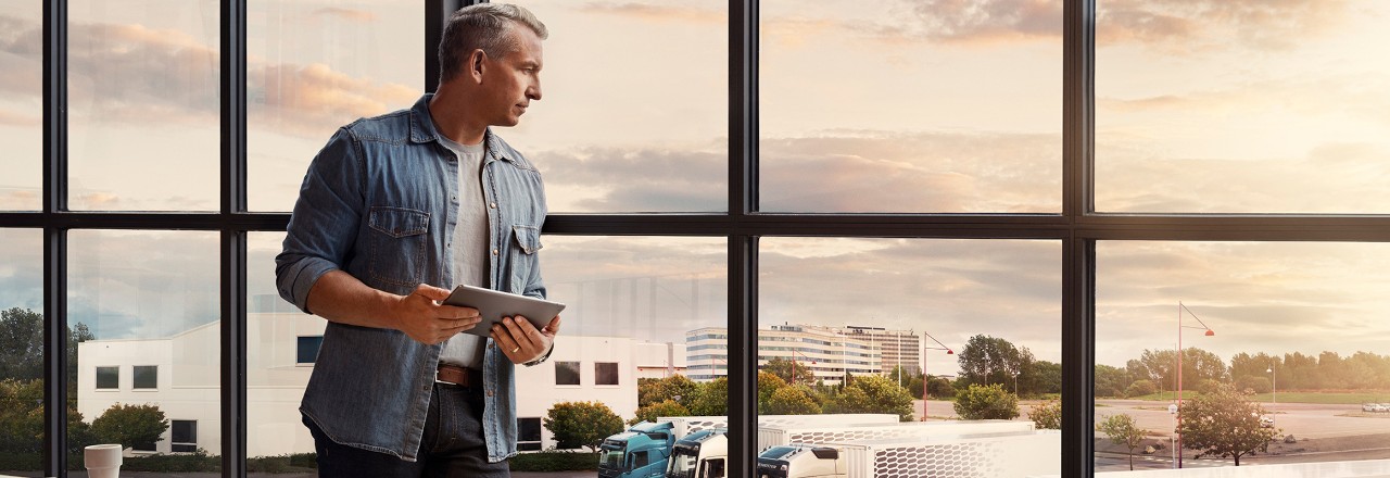 Elinde tablet tutan bir adam pencerenin yanında duruyor ve kamyon filosuna bakıyor